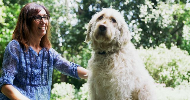 Woman wearing glasses and blue shirt enjoying time with her fluffy dog in a vibrant garden during a sunny day. Ideal for usage in pet care advertisements, gardening catalogs, or lifestyle blogs focused on outdoor activities and companionship.