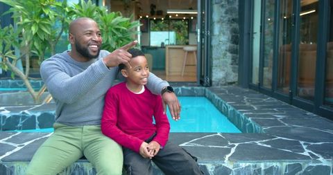 African american father and son sitting by modern pool pointing and smiling, outdoor family moment