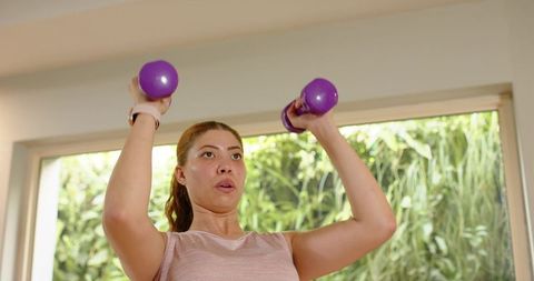 Home shoulder workout: woman lifting dumbbells by window wearing fitness tracker