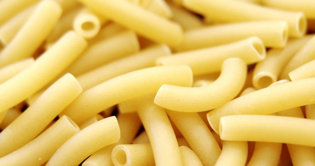 Uncooked macaroni pasta is scattered across the frame, providing a close-up view of the dry, tubular noodles. Ideal for culinary themes, the image captures the simplicity of pasta before it's transformed into a delicious meal.