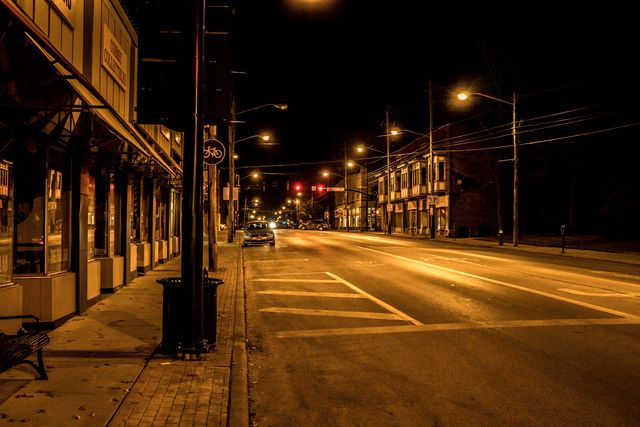 Image shows an empty urban street at night illuminated by streetlights. Quiet atmosphere and empty storefronts create a serene yet isolated feeling. Suitable for projects involving urban scenes, night photography, introspective moments, or illustrating city landscapes at night. Useful for travel blogs, photographic art displays, or aesthetic-focused social media content.