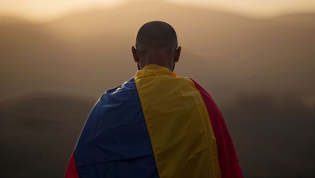 Silhouetted man draped in tricolor flag gazing over layered mountain ridges at sunset