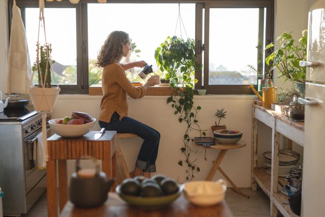 Caucasian woman sitting in a cozy kitchen, pouring coffee while self-isolating and social distancing during quarantine lockdown. The kitchen is filled with natural light and indoor plants, creating a serene atmosphere. This image is perfect for articles or advertisements related to home life, quarantine activities, morning routines, or promoting a relaxed lifestyle.