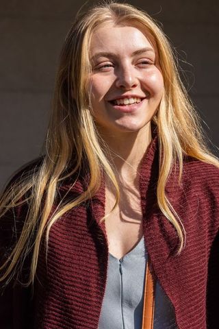 Smiling young blonde woman wearing maroon cardigan and gray zip top in sunlight