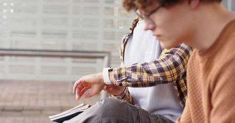 Young diverse commuters checking smartwatch and notebook while waiting at transit shelter