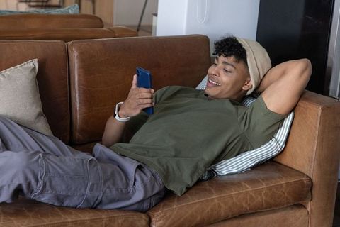 African american man reclining on leather sofa using smartphone, relaxed modern lifestyle