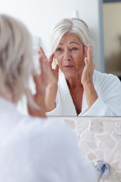 Mature woman with white hair wearing a robe, inspecting her face in a bathroom mirror. Ideal for content related to senior lifestyle, health and beauty routines, skincare products, and self-care practices.