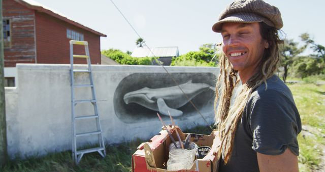 Image of caucasian male artist with dreadlocks holding paints and smiling in front of mural on wall. Freedom, creativity, inclusivity and hobbies concept digitally generated image.