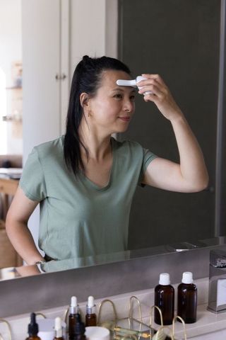 Mature asian woman applying handheld facial device to forehead at bathroom vanity mirror