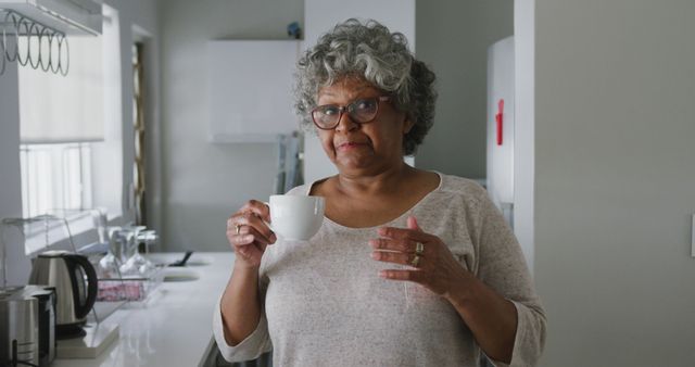 Elderly woman with grey hair and glasses enjoying tea in a modern kitchen. She is casually dressed and appears relaxed and comfortable. This can be useful in articles or advertisements focusing on senior living, healthy lifestyles for the elderly, home life, or family care.