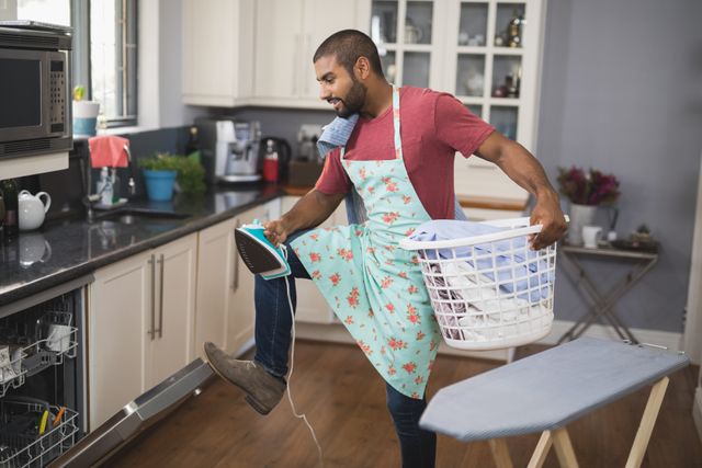 Young man multitasking in kitchen, holding laundry basket and iron while closing dishwasher. Ideal for illustrating domestic life, household chores, and daily routines. Useful for articles, blogs, and advertisements related to home management, cleaning services, and lifestyle tips.