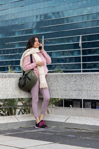 Woman leaning on balustrade calling on smartphone holding olive duffel in pink athleisure