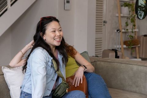 Young women laughing on couch during cozy apartment hangout with plants and luggage