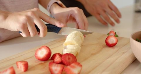 Hands slicing banana into rounds on wooden cutting board with fresh strawberries in home kitchen
