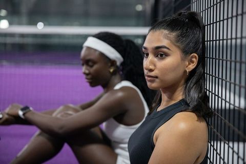 Female athletes resting against court fence checking smartwatch during indoor training