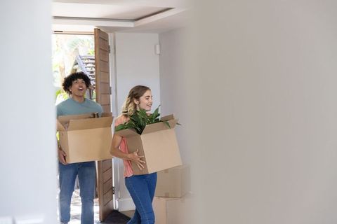 Young couple moving into new home carrying boxes and houseplant through open wooden door