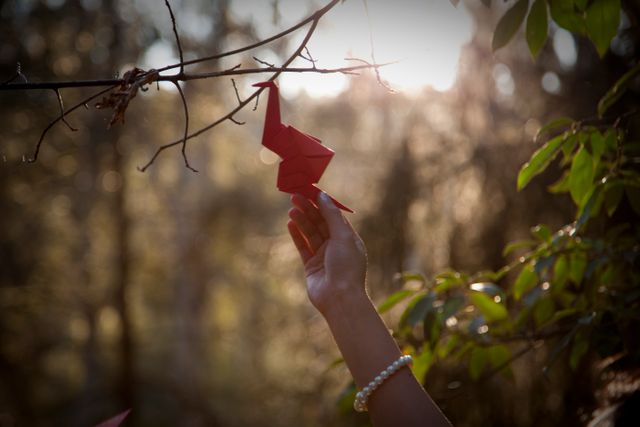 A hand, with a pearl bracelet, is reaching out to hang a red origami crane on a tree branch in a forest. Sunlight filters through the trees, creating a serene atmosphere. This photo can be used to depict concepts related to nature, crafting, calmness, and tranquility. It is suitable for websites about handmade crafts, mindful activities, environmental awareness, and peaceful retreats.