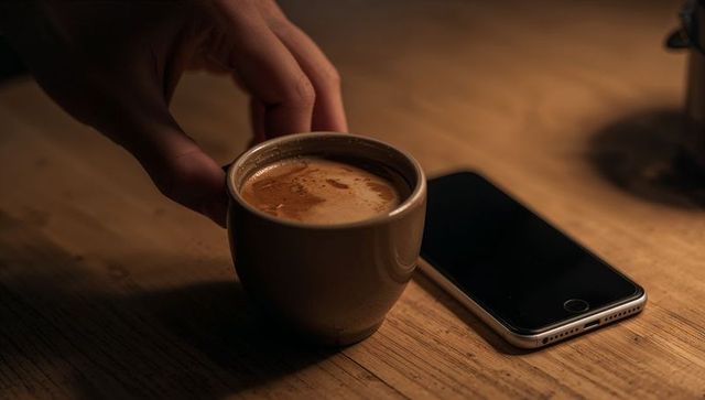 Reaching hand touching espresso cup next to smartphone on rustic wooden table