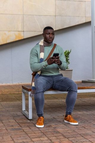 African american man sitting on bench holding smartphone in mint sweater and scarf