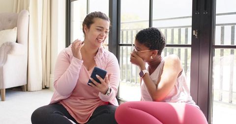Diverse friends sharing smartphone and laughing together in bright living room