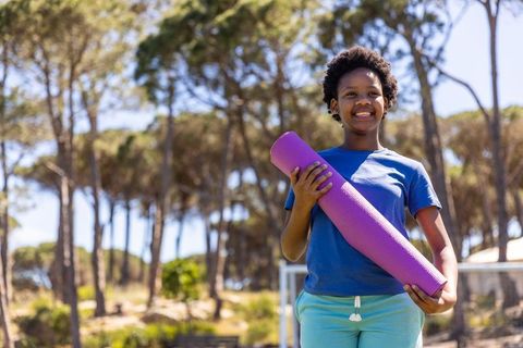 Smiling african american teen holding purple yoga mat in sunny park for outdoor fitness