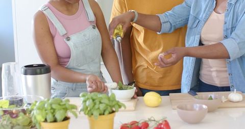 African american friends preparing pesto together in bright kitchen, pouring oil, grinding herbs