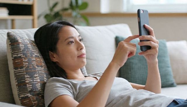 Asian woman reclining on sofa tapping smartphone, relaxing and browsing at home