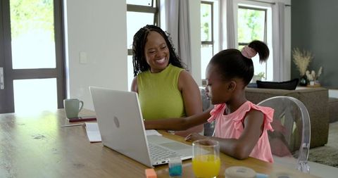 African american mother teaching daughter using laptop at dining table for home learning