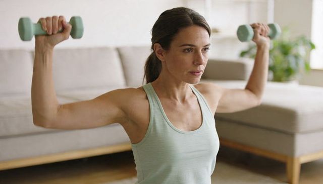 Woman lifting light-green dumbbells during home strength workout in neutral living room