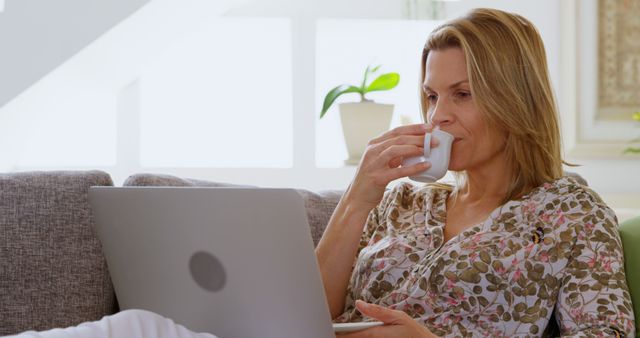 Mature woman in a casual floral shirt is sitting on a couch while drinking coffee and using a laptop. This image is perfect for illustrating concepts related to remote work, freelancing, digital lifestyle, comfort at home, and morning routines. Ideal for blog posts, articles, and advertisements that promote work-from-home culture or comfortable living.