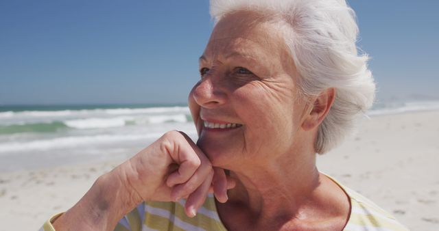 Senior woman smiling at beach on a sunny day with ocean in background. Suitable for use in retirement advertisements, health and wellness campaigns, travel brochures, and lifestyle blogs promoting relaxation.