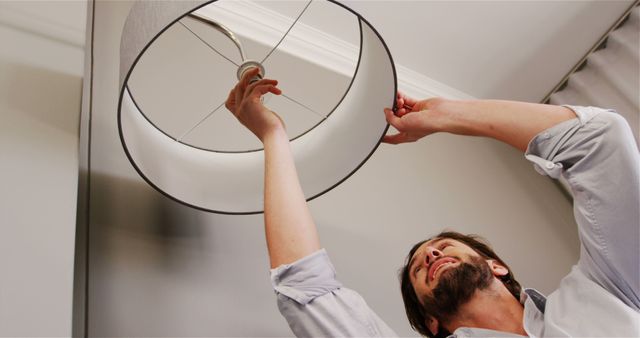 Man adjusting a ceiling light fixture with a grey lampshade. Ideal for use in content related to home improvement, DIY projects, electrical work, handyman services, or repair and maintenance guides.