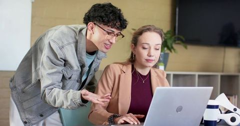 Young colleagues collaborating over laptop in office, coworker leaning and explaining