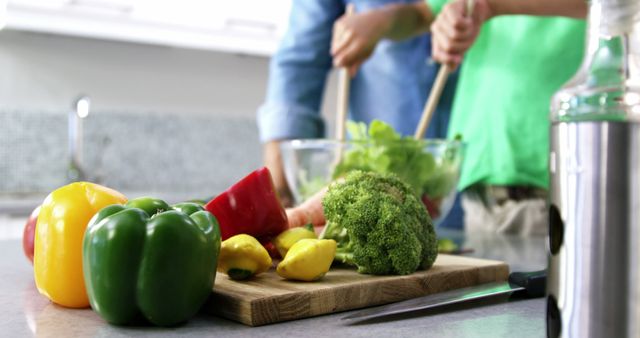 A couple is seen preparing a healthy meal with a variety of fresh vegetables including bell peppers, broccoli, and other assorted produce on a kitchen counter. The individuals are engaged in the process with wooden spoons and bowls. This scene promotes concepts of teamwork in cooking, healthy eating habits, and domestic lifestyle. It can be used for topics related to home cooking, nutritional guides, kitchen equipment, or relationship dynamics.