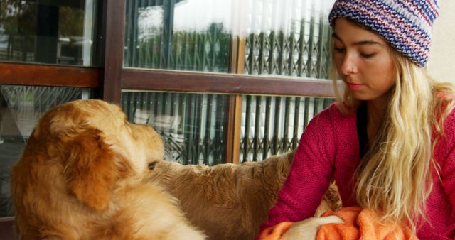Young woman wearing colorful knit hat and pink sweater sitting calmly indoors with her golden retriever puppy. Perfect for illustrating themes of companionship, pet care, leisure time, and the bond between humans and their pets.
