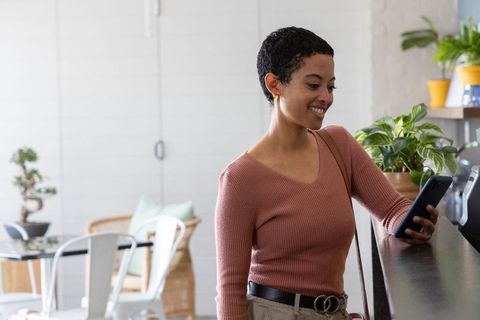 African american woman using smartphone at cafe counter, smiling with plants and sunlight