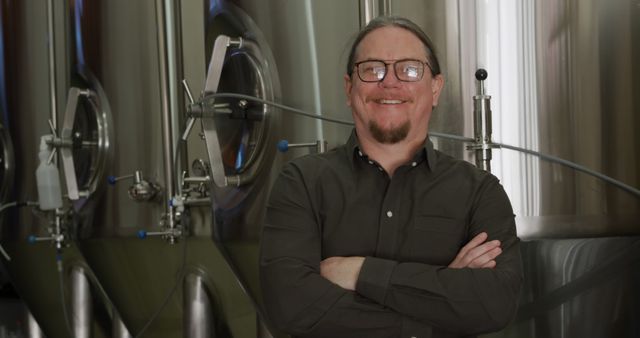 Smiling male brewer standing confidently in front of large metallic vessels used in beer production. Suitable for use in business promotions, beer brewing industry websites, advertisements for brewery tours, and other marketing materials related to brewing and beer production.