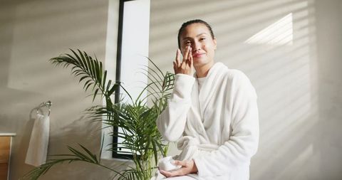 Mid-adult asian woman applying moisturizer in bathrobe by sunlit bathroom window with palm