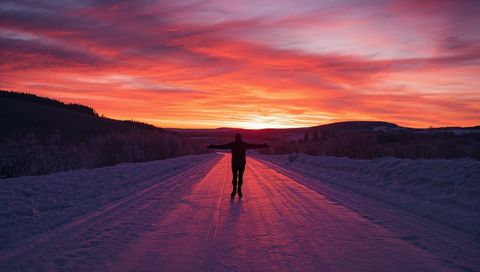 Standing silhouette extending arms on icy road toward fiery winter sunrise over hills