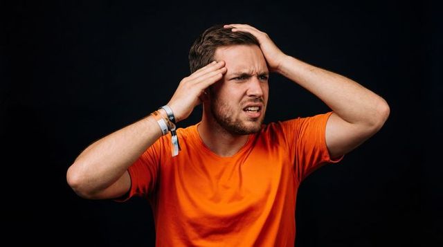 Man holding head looking confused in orange t-shirt with wristbands on black background