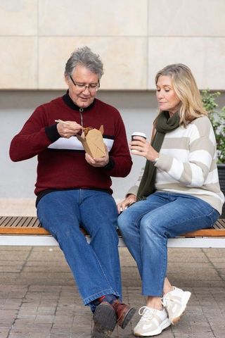Older couple eating takeout noodles on bench outdoors, sharing coffee and conversation