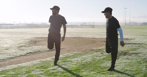 Two Teammates Stretching on Dew-Covered Baseball Infield at Sunrise, Backlit Warmup
