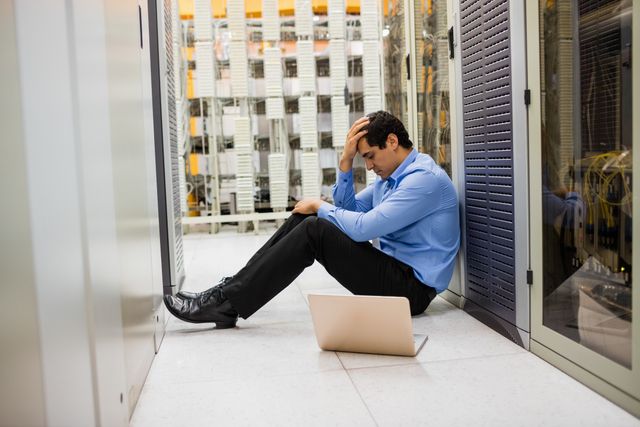 Technician sitting on floor in server room hallway, looking stressed and frustrated. Laptop open beside him, indicating technical issues or troubleshooting problems. Ideal for illustrating work stress, IT challenges, or data center environments. Useful for articles on technology, business, or mental health in the workplace.