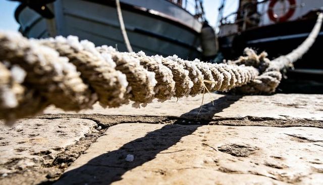 Fraying mooring rope coated with salt crystals lying diagonally on sunlit stone quay, boats nearby