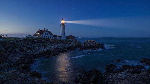 Lighthouse beacon casting beam across rocky coastline at twilight