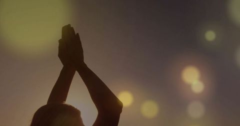 Woman silhouette raising hands and pressing palms together at sunset with bokeh halo