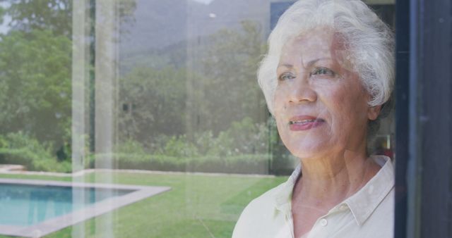 Happy senior biracial woman standing by window at home smiling, with garden and pool in background. Senior lifestyle, tranquility and domestic life.
