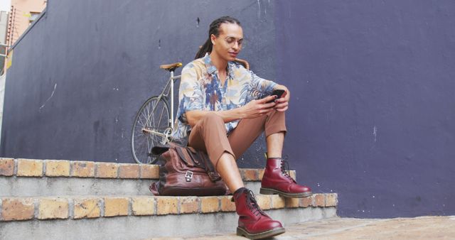 A young man with dreadlocks sitting on outdoor stairs, checking his phone. He is dressed in casual, stylish attire, featuring a patterned shirt and red boots. A bicycle is placed against a wall in the background, adding to the urban aesthetic. This can be used for communications, fashion, lifestyle blogs, urban living themes, and modern technology use illustrations.