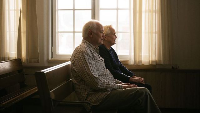 Senior couple sitting on wooden bench gazing through windowpane in quiet sunlit interior