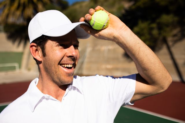 Side view of a happy Caucasian man wearing tennis whites playing tennis on a sunny day, smiling at camera and holding his cap and a tennis ball.